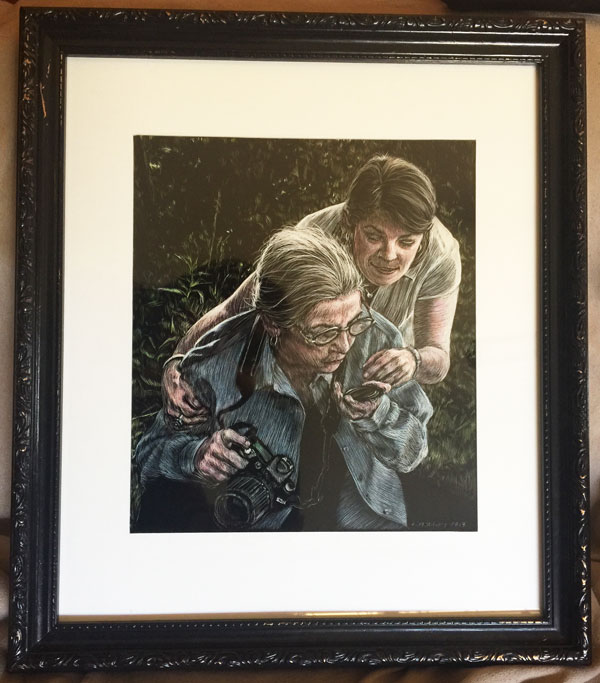 Mother the photographer blowing dust of her lens cap as her daughter leans over to watch rendered in scratchborad and color ink by Lori McAdams in a black carved frame