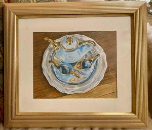 Gold framed drawing of a blue and gold blue porcelain teaset on a silver platter against a wooden table.