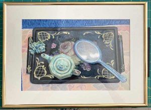 Black laquer tray with celedon teapot and flowers and blue lucite mirror on a pale floral place mat in golden strip frame.